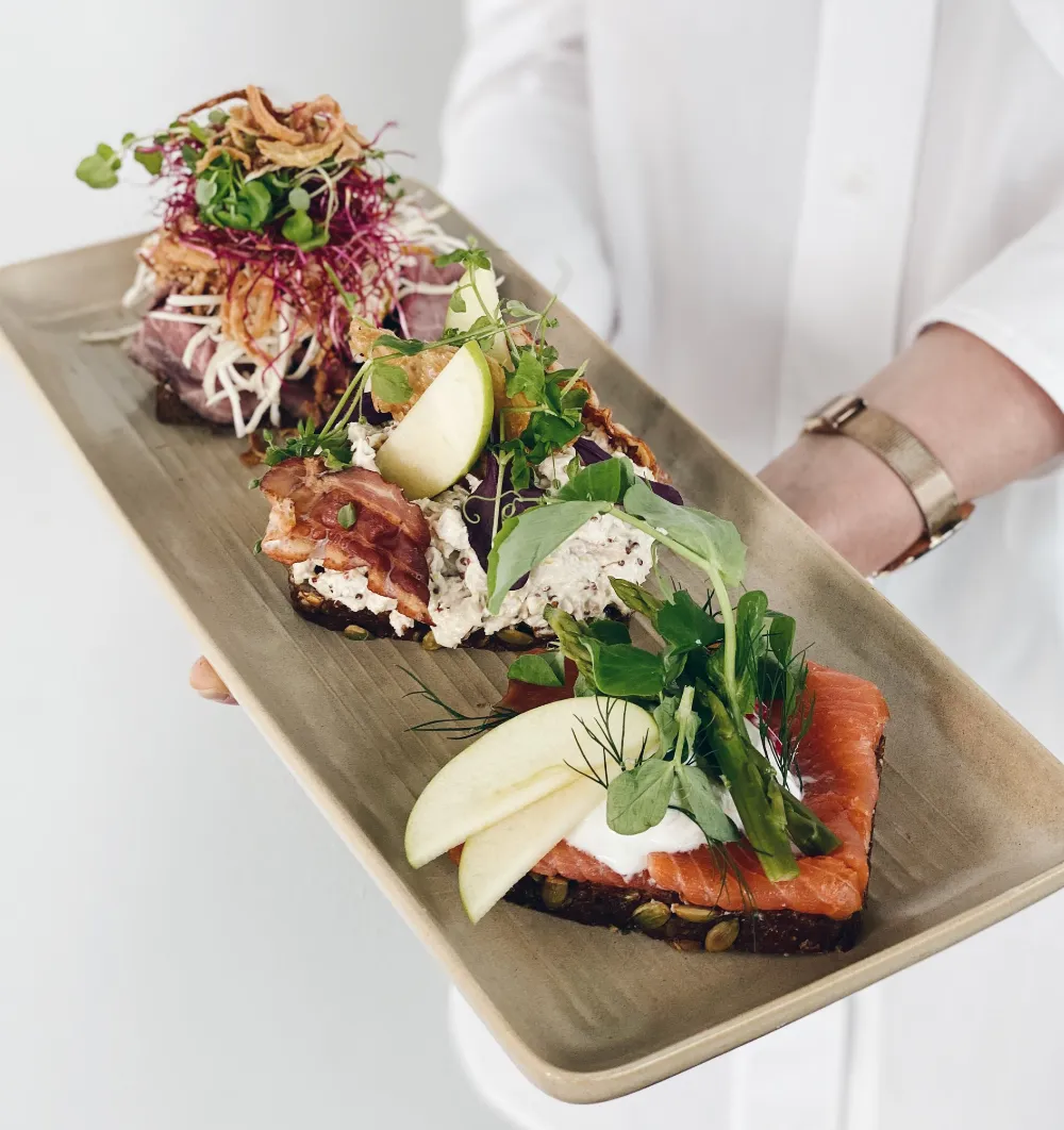 Gourmet smørrebrød med laks, æble og urter fra frokostordning Malling serveret på aflang stentøjstallerken.